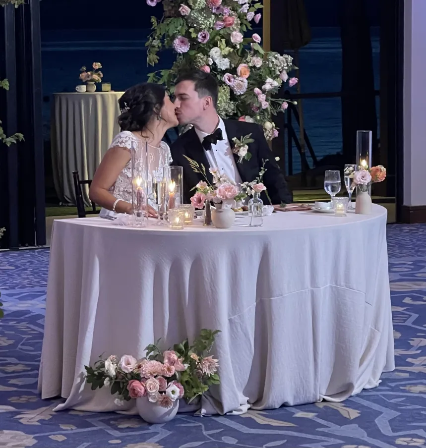Sheila and Evan kissing at their sweetheart table, candles and pink roses in front of them and the water behind