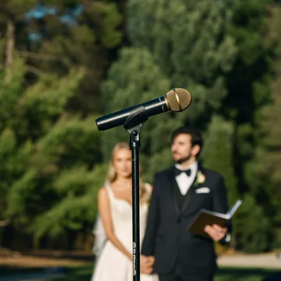 Microphone on a stand at an outdoor wedding ceremony, the couple softly out of focus behind it